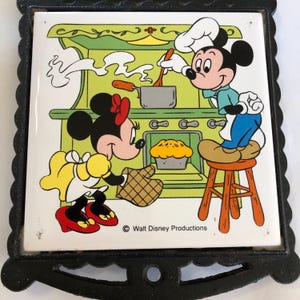 May include: A black and white ceramic tile trivet featuring Mickey and Minnie Mouse in a kitchen setting. Mickey is wearing a chef's hat and apron, stirring a pot on the stove. Minnie is wearing a yellow dress and red shoes, holding a potholder. The tile has the text "Walt Disney Productions" at the bottom.