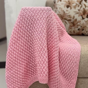 Puede incluir: Una suave manta rosa tejida sobre un sofá de tonos neutros. La manta tiene una apariencia texturizada y tejida y está doblada para revelar su grosor. Un cojín con estampado floral es visible en el fondo.