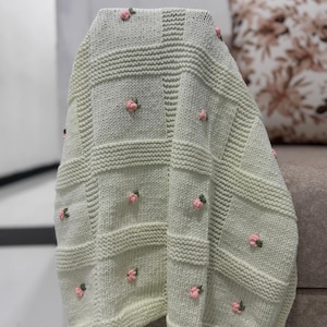 May include: A light green knitted blanket with a square pattern and embroidered pink roses. The blanket is draped over a surface, showcasing its texture and floral details. The blanket is a cozy home decor item.