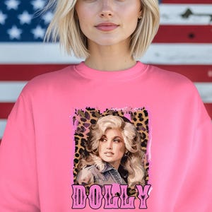 May include: A bright pink sweatshirt featuring a portrait of a blonde woman with the word "DOLLY" in pink script. The portrait is set against a leopard print background. The background is an American flag.