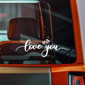 Puede incluir: Calcomanía de vinilo blanco con las palabras "love you" en fuente cursiva, acompañada de dos pequeños símbolos de corazón. La calcomanía está aplicada en la ventana de un vehículo naranja, con el espejo lateral visible.