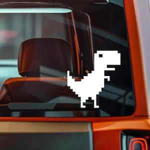 May include: A white pixelated dinosaur decal, reminiscent of classic video game graphics, is affixed to a vehicle's window. The dinosaur is in a standing pose, with its tail and head clearly defined. The decal is set against the backdrop of a car's interior and exterior.