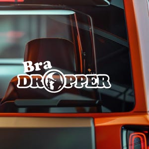 May include: White vinyl decal with the words "Bra DROPPER" in a bold, outlined font. The word "DROPPER" features a graphic of a person. The decal is displayed on a vehicle's rear window, with an orange vehicle frame visible.
