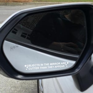 Könnte beinhalten: Weißer Seitenspiegel eines Autos mit schwarzem Rahmen. Der Spiegel zeigt den Text "OBJECTS IN THE MIRROR ARE CUTTER THAN THEY APPEAR" in Weiß, mit Herzsymbolen, auf weißem Hintergrund.