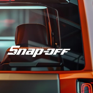 May include: White "Snap-Off" decal on a vehicle window. The decal is in a bold, sans-serif font. The vehicle is orange and black, with a side mirror visible. The background is blurred.