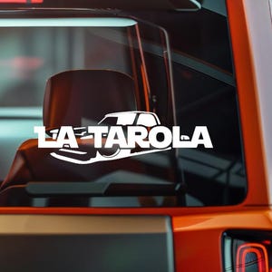 May include: White vinyl decal featuring the text "LA TAROLA" with a stylized car silhouette. The decal is applied to the rear window of an orange vehicle, with the interior visible through the glass.