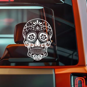May include: White decorative sugar skull decal with floral and ornamental details. The decal is applied to a vehicle window, showcasing the intricate design of the skull, perfect for Day of the Dead celebrations.