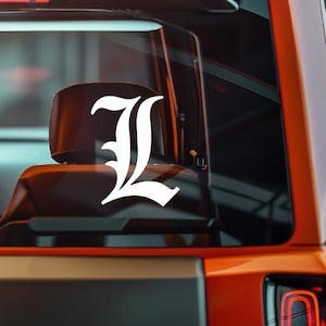 Op de afbeelding: Witte vinyl sticker van de letter "L" in een gotisch lettertype, bevestigd op de achterruit van een voertuig. De sticker valt op tegen het donkere interieur en de oranje buitenkant van het voertuig.
