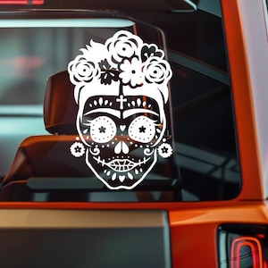 May include: White vinyl decal featuring a stylized sugar skull with a floral crown. The design includes intricate details, such as flower accents, dotted patterns, and a cross. The decal is likely for a car window or similar surface.