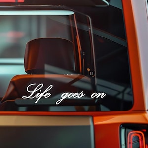 以下が含まれることがあります： オレンジ色の車のリアウィンドウに貼られた、筆記体フォントで「Life goes on」と書かれた白いビニールデカール。 デカールは中央に配置され、暗いインテリアに対して見えます。