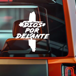 Könnte beinhalten: Weißer Vinyl-Aufkleber mit dem Schriftzug "DIOS POR DELANTE" in einer stilisierten Schriftart, begleitet von einem Kreuz-Design. Der Aufkleber ist auf der Heckscheibe eines orangefarbenen Fahrzeugs angebracht.