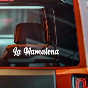 May include: White vinyl decal with the text "La Mamalona" in a cursive font, applied to a vehicle's rear window. The decal is visible against the dark interior and the orange exterior of the vehicle.
