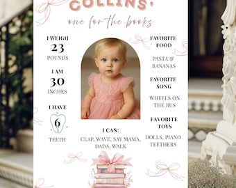 May include: A white milestone birthday sign featuring a central photo of a child in a pink dress, with text including 'COLLINS', 'one for the books', '23 POUNDS', '30 INCHES', '6 TEETH', and lists of favourite foods, songs, toys, and abilities.