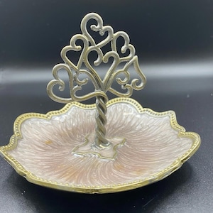 May include: A decorative trinket dish with a heart-shaped tree design. The dish has a light pink and gold-colored base with a twisted stem and a tree-like structure made of hearts. The dish is approximately 6 inches in diameter.