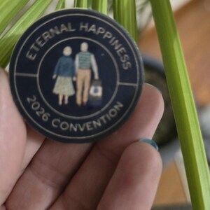 May include: A navy blue button with gold lettering and a graphic of two people holding hands. The text on the button reads "ETERNAL HAPPINESS 2026 CONVENTION". The button is round and has a gold border.