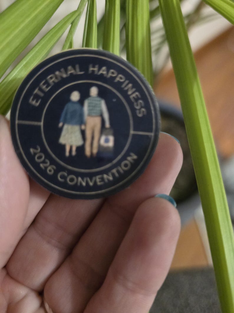May include: A navy blue button with gold lettering and a graphic of two people holding hands. The text on the button reads "ETERNAL HAPPINESS 2026 CONVENTION". The button is round and has a gold border.
