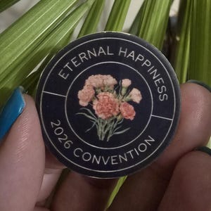 May include: A round, navy blue button with the words "ETERNAL HAPPINESS 2026 CONVENTION" encircling a floral design. The flowers are a mix of pink and peach. The button is held between fingers against a background of green leaves.