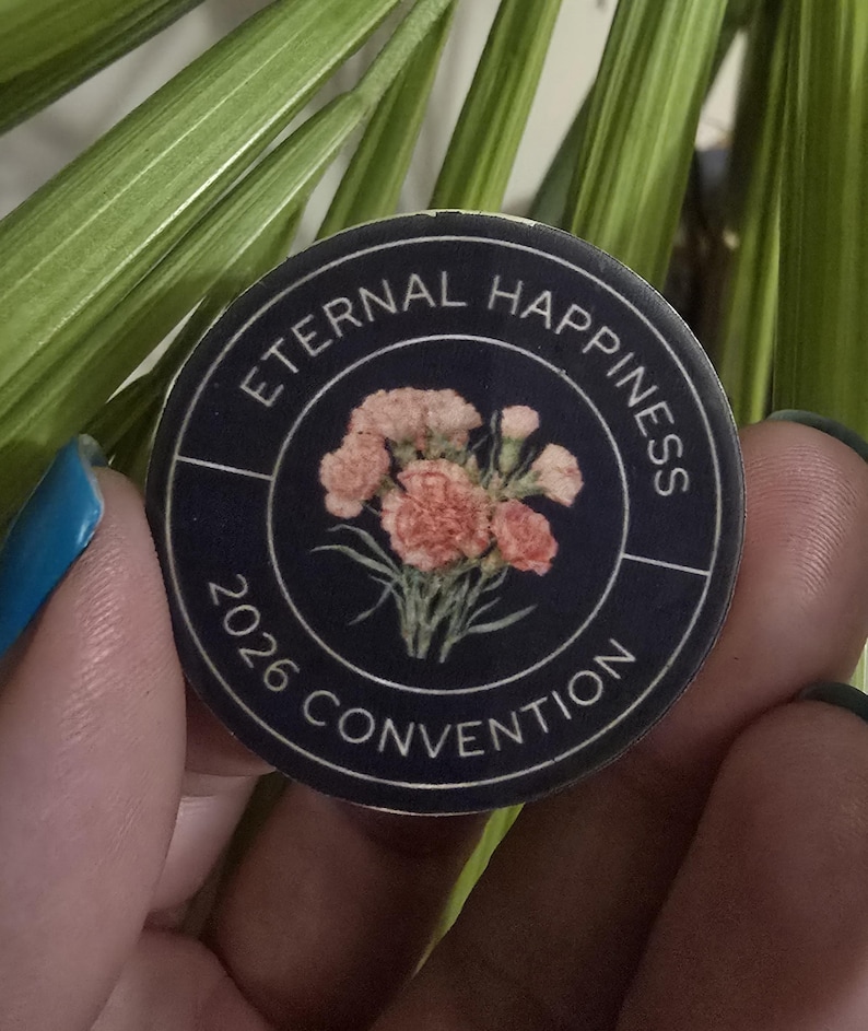 May include: A round, navy blue button with the words "ETERNAL HAPPINESS 2026 CONVENTION" encircling a floral design. The flowers are a mix of pink and peach. The button is held between fingers against a background of green leaves.