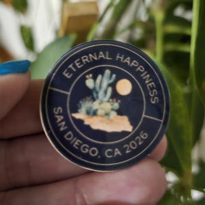 May include: A round, navy blue pin with the words "Eternal Happiness" and "San Diego, CA 2026" in white. The pin features a desert landscape with cacti and a full moon.