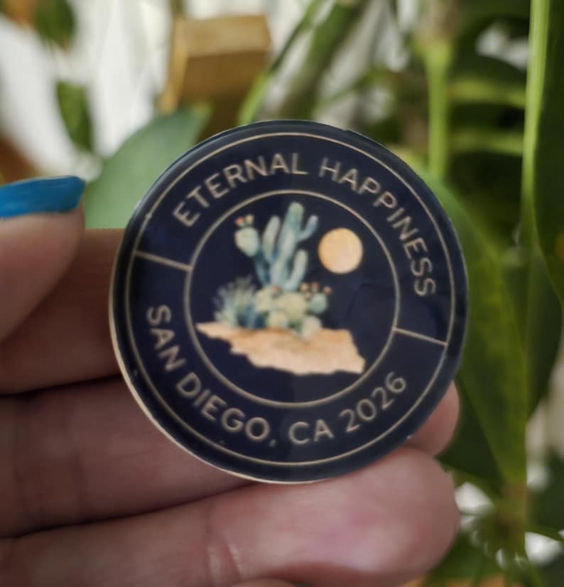 May include: A round, navy blue pin with the words "Eternal Happiness" and "San Diego, CA 2026" in white. The pin features a desert landscape with cacti and a full moon.