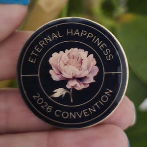 May include: A round, black pin with gold trim featuring the text "ETERNAL HAPPINESS 2026 CONVENTION." A pink and white peony flower is in the center of the pin. The pin is held in a hand.