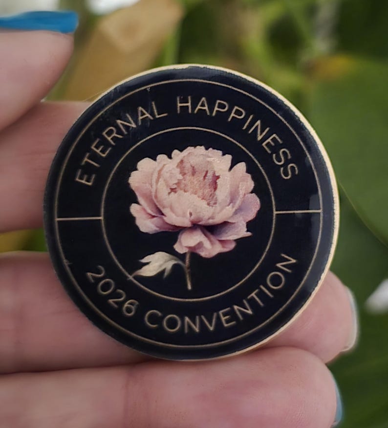 May include: A round, black pin with gold trim featuring the text "ETERNAL HAPPINESS 2026 CONVENTION." A pink and white peony flower is in the center of the pin. The pin is held in a hand.