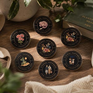 May include: A selection of black and gold enamel pins with various designs, including flowers and landscapes. Each pin is inscribed with "Eternal Happiness 2026 Convention". The pins are displayed on a wooden surface, with a book and a mug nearby.
