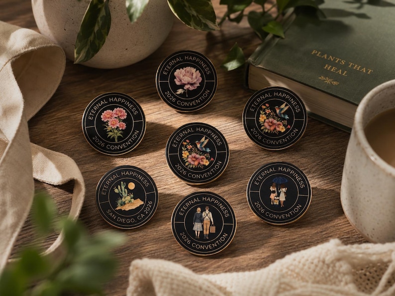 May include: A collection of black and gold enamel pins with floral and scenic designs. Each pin features the text "Eternal Happiness 2026 Convention". The pins are arranged on a wooden surface, alongside a book and a mug.
