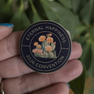May include: A round, navy blue button with the words "Eternal Happiness" and "2026 Convention" in gold lettering. The center features a watercolor illustration of cacti and orange flowers.