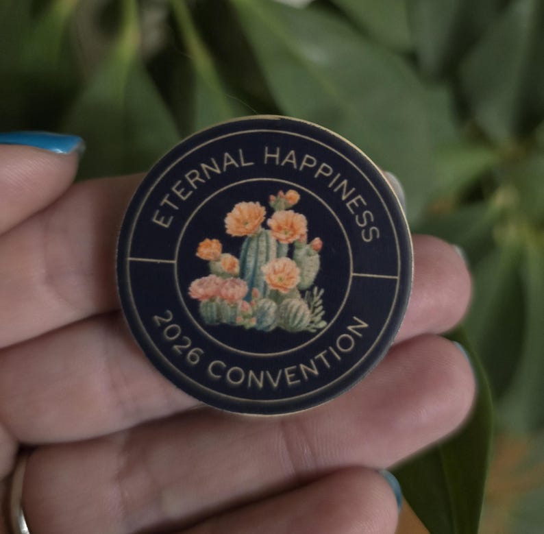 May include: A round, navy blue button with the words "Eternal Happiness" and "2026 Convention" in gold lettering. The center features a watercolor illustration of cacti and orange flowers.