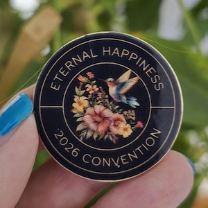 May include: A round, black button with gold trim featuring the text "ETERNAL HAPPINESS 2026 CONVENTION." The button's center showcases a hummingbird in flight above a bouquet of flowers in shades of pink, yellow, and orange.
