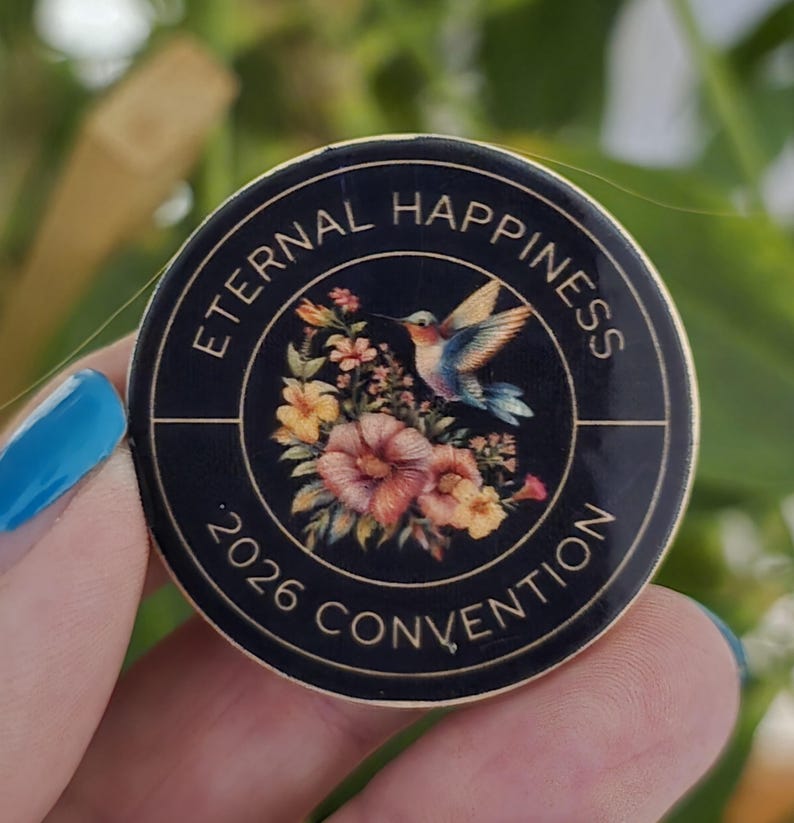 May include: A round, black button with gold trim featuring the text "ETERNAL HAPPINESS 2026 CONVENTION." The button's center showcases a hummingbird in flight above a bouquet of flowers in shades of pink, yellow, and orange.
