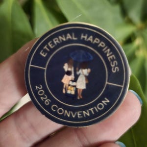 May include: A navy blue round pin with the words "Eternal Happiness 2026 Convention" in white. The pin features an illustration of two figures under an umbrella. The pin is held in a hand.