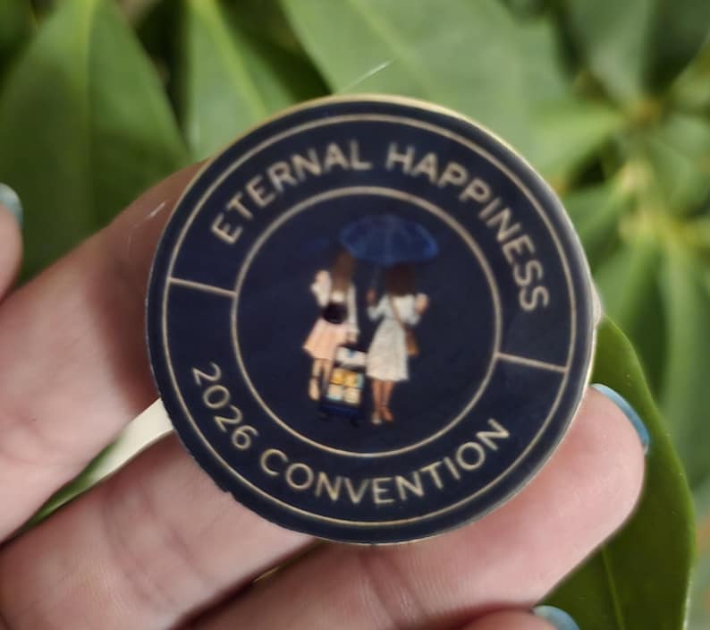May include: A navy blue round pin with the words "Eternal Happiness 2026 Convention" in white. The pin features an illustration of two figures under an umbrella. The pin is held in a hand.