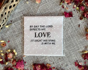Könnte beinhalten: Eine quadratische, weiße Karte mit den Worten "BY DAY THE LORD DIRECTS HIS LOVE AT NIGHT HIS SONG IS WITH ME" in Schwarz. Die Karte hat ein Tupfenmuster und ist von getrockneten Blumen umgeben.