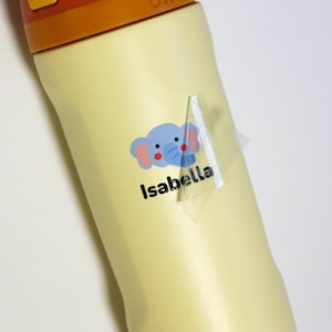 May include: A light yellow bottle with a sticker featuring a blue elephant with pink ears and the name "Isabella" in black text. The sticker is partially peeled off.