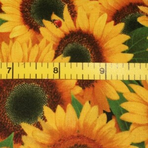 May include: A close-up of a fabric with a sunflower pattern. The fabric is yellow, brown, and green. A yellow measuring tape is placed across the fabric, with the numbers 7, 8, 9, and 10 visible.