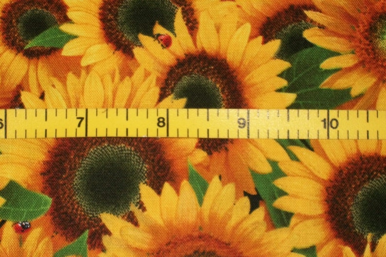 May include: A close-up of a fabric with a sunflower pattern. The fabric is yellow, brown, and green. A yellow measuring tape is placed across the fabric, with the numbers 7, 8, 9, and 10 visible.