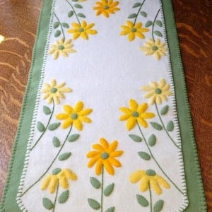 May include: A white felt table runner with a green border, featuring yellow daisy appliqués with green stems and leaves. The design includes the text "©2018 Cath's Pennies Designs". The runner is placed on a wooden table.