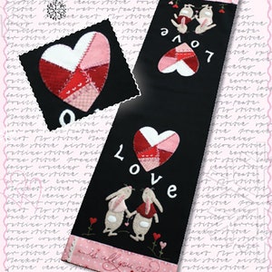 May include: A black fabric wall hanging with a pink and red patchwork heart, two bunnies holding hands, and the words "Love is all you need".