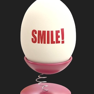 May include: A white egg with the word "SMILE!" in red lettering, resting in a pink egg cup with a spring base. The egg cup and base are a matching shade of pink, set against a black background.