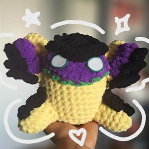 May include: A handmade crochet plush toy in the shape of a creature with yellow, purple, black, and green accents. The toy has large, round white eyes and is embellished with white doodle art. The plush is held in a hand.