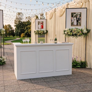 Può includere: Allestimento per ricevimento di nozze all'aperto con bar bianco, foto incorniciate, composizioni floreali e luci a corda. Il bar ha un design semplice ed è posizionato su un patio in pietra. Lo sfondo presenta un fondale in tessuto drappeggiato.
