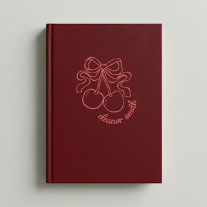 Op de afbeelding: Een bordeauxrood notitieboek met een roze kersenillustratie en de naam "eleanor smith". De omslag toont een paar kersen vastgebonden met een strik. Het notitieboek heeft een strak, minimalistisch ontwerp.