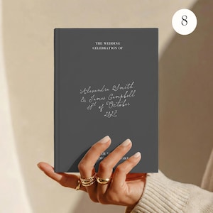 May include: A dark grey hardcover book with white text that reads THE WEDDING CELEBRATION OF Alexandra Smith & James Campbell 10th of October 2027. The cover features a clean, matte finish with elegant, handwritten-style script for the names and date.