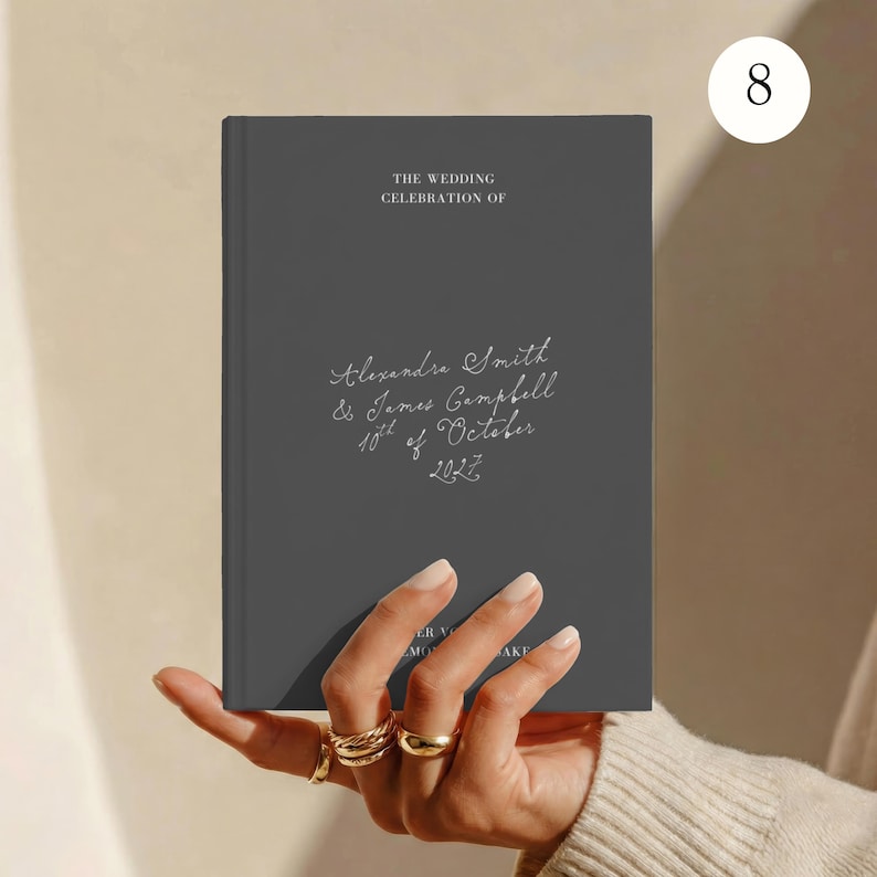 May include: A dark grey hardcover book with white text that reads THE WEDDING CELEBRATION OF Alexandra Smith & James Campbell 10th of October 2027. The cover features a clean, matte finish with elegant, handwritten-style script for the names and date.