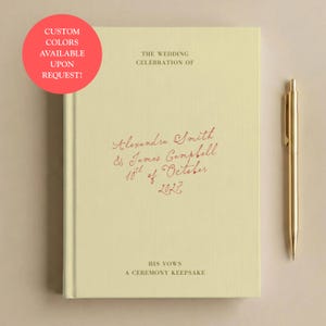 May include: A pale yellow hardback book with the text THE WEDDING CELEBRATION OF, Alexandra Smith & James Campbell 10th of October 2027, HIS VOWS, A CEREMONY KEEPSAKE printed on the cover. A gold-coloured metal pen is positioned to the right of the book.