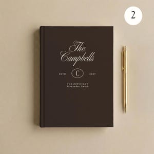 May include: A dark brown hardcover book with white text reading The Campbells, ESTD 2027, and THE OFFICIANT Alexander Smith, featuring a central oval with the letter C. A gold-colored metal pen is positioned vertically to the right of the book.