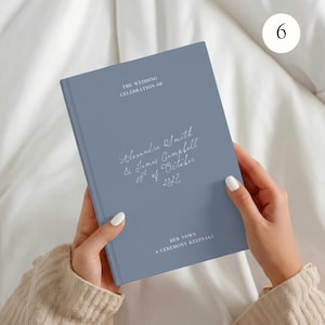 May include: A dusty blue hardback book with white text that reads THE WEDDING CELEBRATION OF, Alexandre Smith & James Campbell 10th of October 2027, and HER VOWS A CEREMONY KEEPSAKE. The book has a smooth matte finish and a rectangular shape.