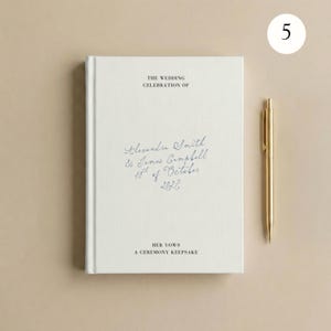 May include: A white hardback book with the text THE WEDDING CELEBRATION OF, Alexandra Smith & James Campbell 10th of October 2027, HER VOWS A CEREMONY KEEPSAKE printed on the cover, accompanied by a gold-coloured metal pen placed to the right.